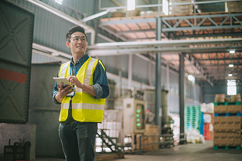 Warehouse worker in safety vest holding tablet, inspecting inventory in large industrial storage facility.