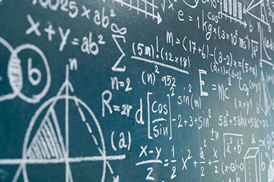 Close-up of a chalkboard covered with handwritten mathematical equations, symbols, and diagrams, including algebraic expressions, geometric shapes, and formulas written in white chalk.