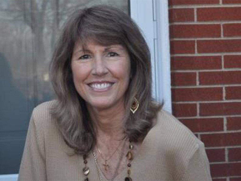 Smiling woman with shoulder-length brown hair wearing a beige sweater and long beaded necklace, sitting in front of a brick wall and window.