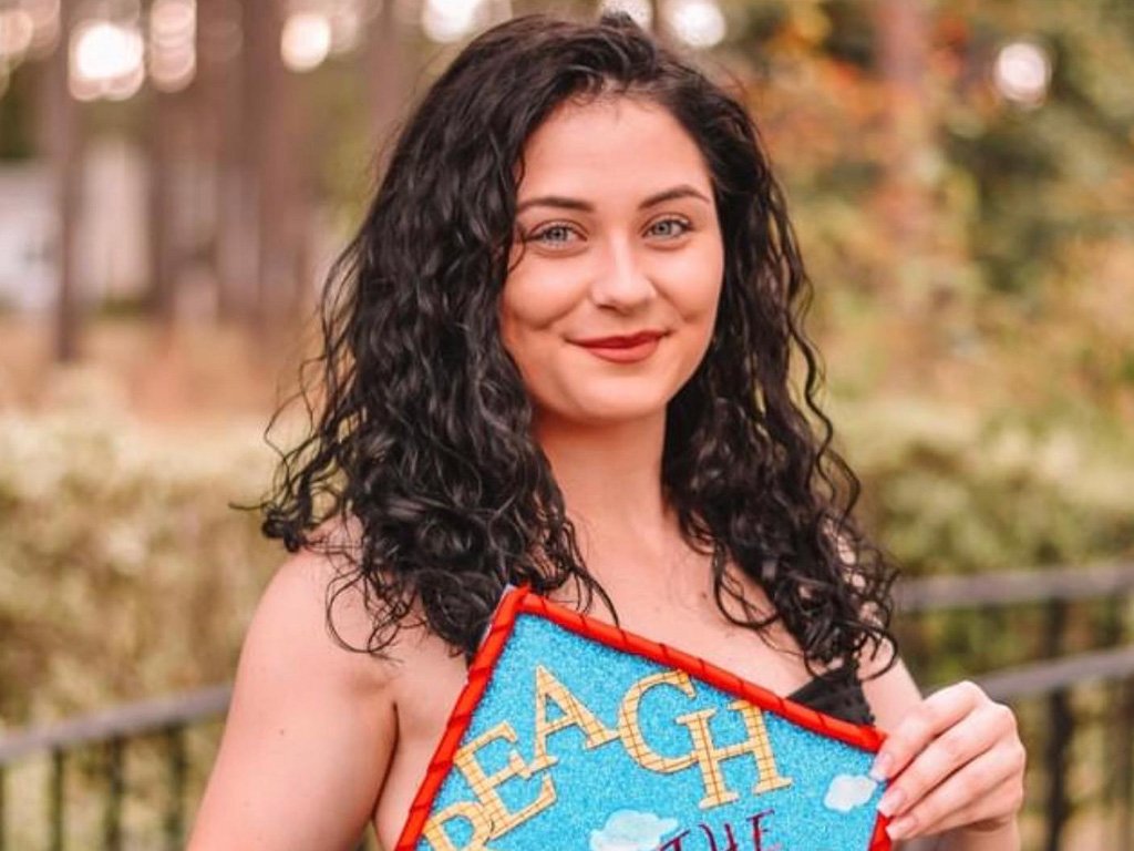 Smiling woman with curly dark hair in a black dress holding a decorated graduation cap that reads ‘Reach for the Sky’ with clouds and a red bow.