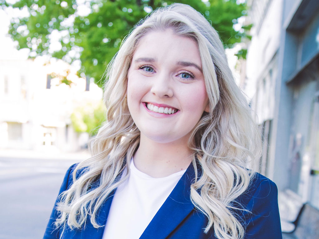 Smiling woman with long blonde hair wearing a blue blazer and white top, standing outdoors on a city street.