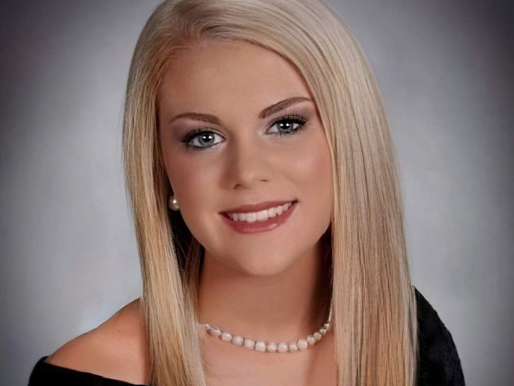 Smiling young woman with straight blonde hair wearing a black off-shoulder top, pearl necklace, and earrings, posed against a gray background.